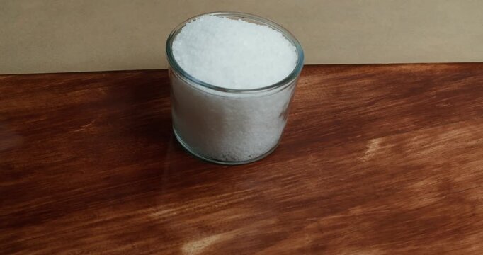 Close up of a chef hand placing a glass filled with coarse sea salt in the center of a wooden table. Natural sea salt is prepared as a seasoning ingredient for homemade cooking in a cozy kitchen.
