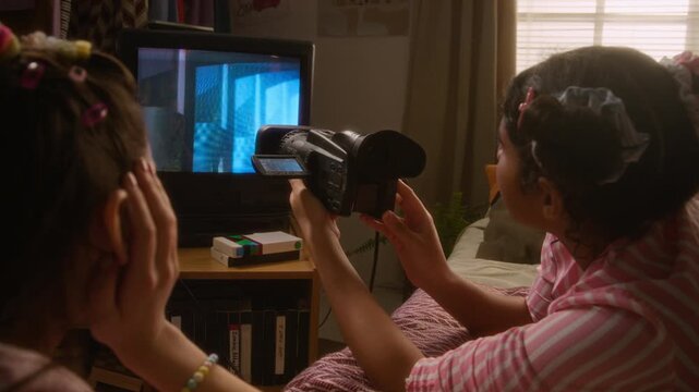 Two diverse teenagers lying on bed, holding retro video camcorder, trying out creative effects on CRT television screen. Nostalgic marketing, retro technology promotions, or youth lifestyle content