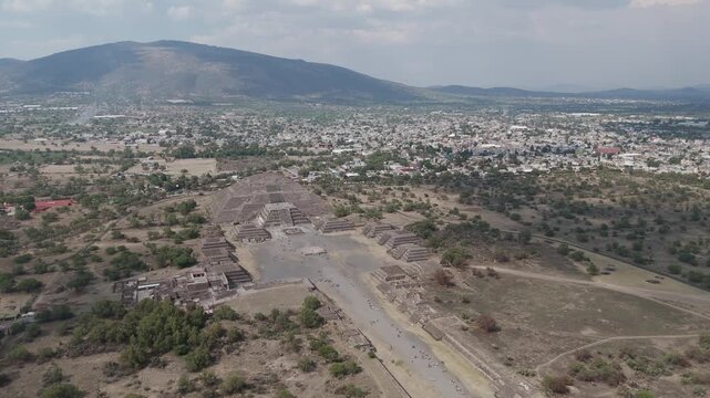 Sweeping aerial perspective of the ancient teotihuacan pyramids, featuring the moon pyramid and the avenue of the dead with the modern city and mountains visible in the distant background