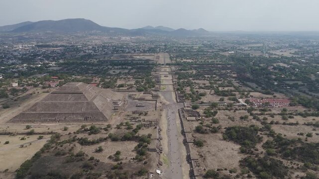 Aerial view flying backwards over the avenue of the dead at the ancient teotihuacan archeological site, revealing the pyramid of the moon with mountains in the background