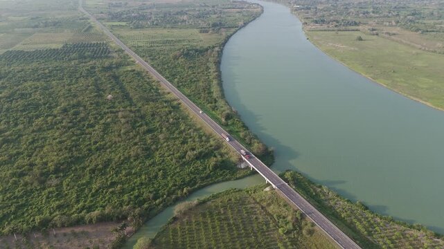 Elevated shot of a long straight road running through a lush green landscape with palm tree plantations and dense forest, with cars and trucks crossing a bridge over curving river in Tuxpan Mexico
