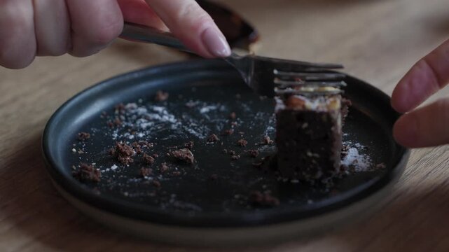 Hand uses fork to cut into a piece of chocolate nut cake on a dark ceramic plate with crumbs