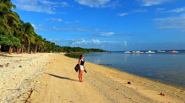 A woman strolls along a picturesque beach with palm trees and calm waters under a bright blue sky.