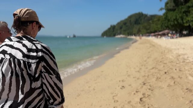 A woman in a zebra-print cover-up and cap walks along Koh Yao Yai's golden sand beach, framed by turquoise sea, palm trees, and distant hills under bright, natural daylight.