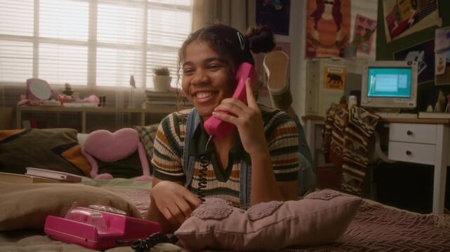 Black teenage high school girl lying on bed, dialing and talking on pink retro telephone while laughing. Footage perfect for nostalgia marketing, telecommunications ads, or youth lifestyle campaigns