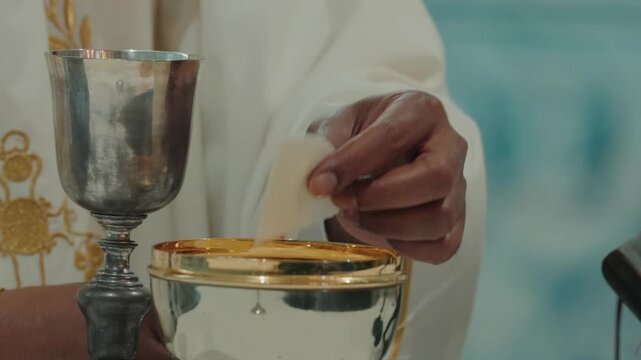 Priest hands place a communion host into a golden ciborium beside a silver chalice during mass