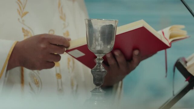 Priest in white vestments holds a silver chalice and red missal during the celebration of Catholic mass