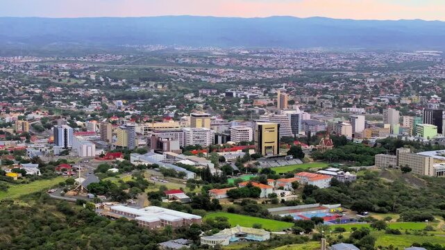 Elevated aerial of Windhoek's downtown and suburbs seen from the surrounding green hills. The capital city spreads across a wide valley ringed by rugged highland mountains under an open sky.