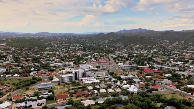 Aerial flight over Windhoek's neighbourhoods and main roads with the city framed by rugged hills. The capital city spreads across a wide valley ringed by rugged highland mountains under an open sky.