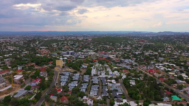 Drone flight over the residential suburbs and rooftops of Windhoek, the capital of Namibia, with distant hills under a cloudy sky. A sprawling capital ringed by highland mountains.