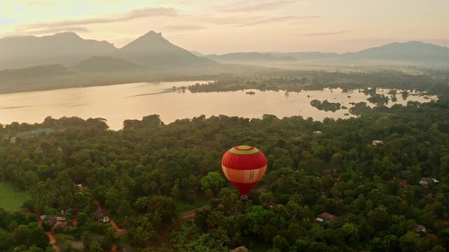 Aerial view of a red and yellow hot air balloon above jungle and a flooded plain at dawn, distant hills bathed in soft morning light.