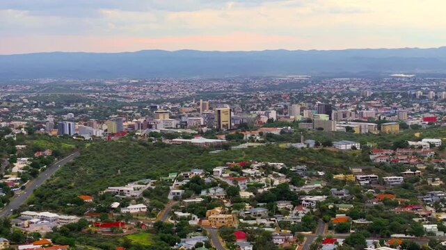 Drone view of central Windhoek with high-rise office buildings and the city skyline backed by mountains at golden light. A sprawling capital ringed by highland mountains.
