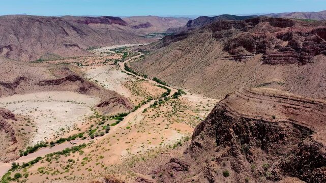 Aerial view of a dramatic switchback pass descending into a Namibian canyon, a green ribbon of riverbed vegetation winding along the gorge floor.
