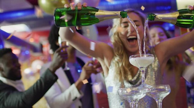 Woman lifting two bottles and pouring into tower at event, foaming while guests filming, cheering
