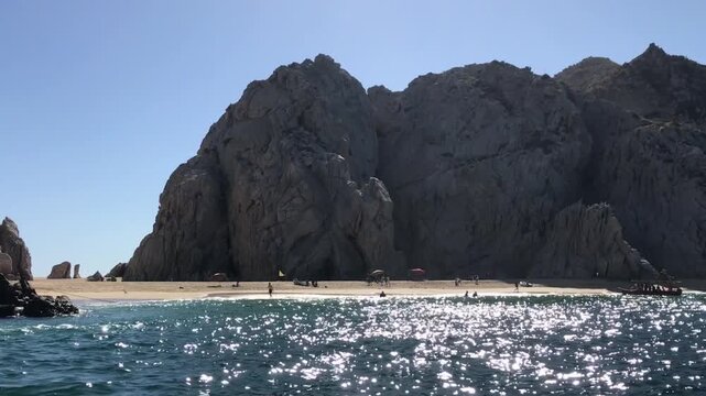 Coastal view of towering granite cliffs rising above a sandy beach and sparkling ocean water, showcasing the dramatic shoreline of Cabo San Lucas, Mexico