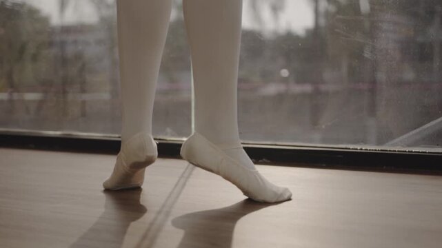 Low section of ballerina legs in white stockings and pointe shoes performing en pointe ballet movements near window inside dance studio
