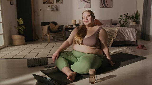 Portrait of plus-size young adult Caucasian woman sitting on fitness mat and smiling at camera. Footage is ideal for body positivity campaigns, home workout apps, and mental wellbeing marketing