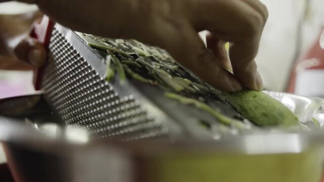 Close up of person grating fresh zucchini with box grater into bowl. Homemade food preparation featuring vegetable ingredient, cooking technique and meal preparation.