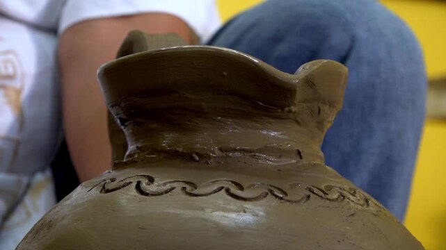 Close up of artisan decorating handmade clay pot with carved pattern before firing. Traditional pottery craft showcasing ceramic artistry, skilled workmanship and cultural heritage