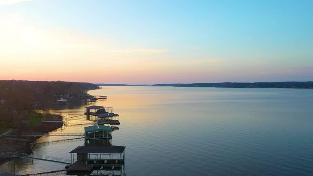 Sunset drone shot near Branson, Missouri, with residential lake shoreline, private boat houses, wooded hills, calm reservoir water, and soft golden reflections in peaceful dusk light.