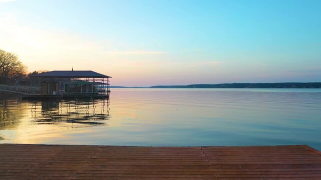 Drone aerial over Branson, Missouri at sunset, with calm Ozark lake water, wooded shoreline, waterfront homes, covered boat docks, pastel sky, and golden reflections across the reservoir.