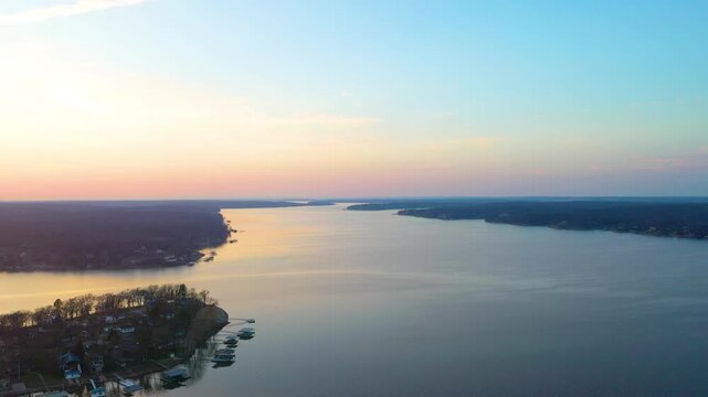 High drone view above a Branson, Missouri lake at dusk, looking across calm water, wooded Ozark shoreline, docked boat houses, lakefront homes, and soft sunset colors over the reservoir.