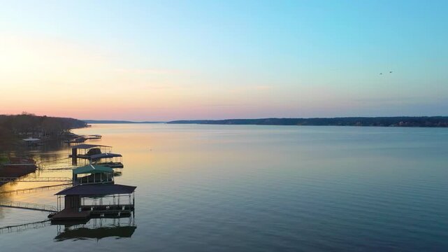 Wide aerial view of Dale Hollow Lake in Tennessee, showing quiet docks, lakeside homes, tree-covered shores, pastel sunset light, and calm open water stretching toward the horizon.