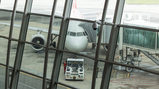 Airplane parking at gate with pushback tractor and jet bridge seen through airport window