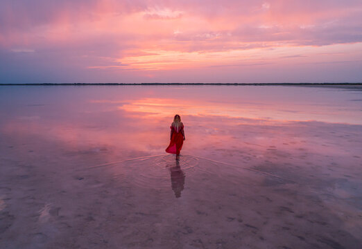 Aerial of a young woman in red dress walking in the water of a unique pink salt lake. Sunset at lake Bursol with beautiful reflections on calm water surface. Stunning scenery