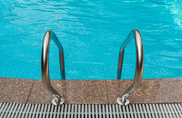 A close-up view of the stainless steel handrails of a swimming pool. The handrails are secured to the deck, which is made of large, brown tiles. The pool is filled with sparkling blue water. © Evgenii Bakhchev