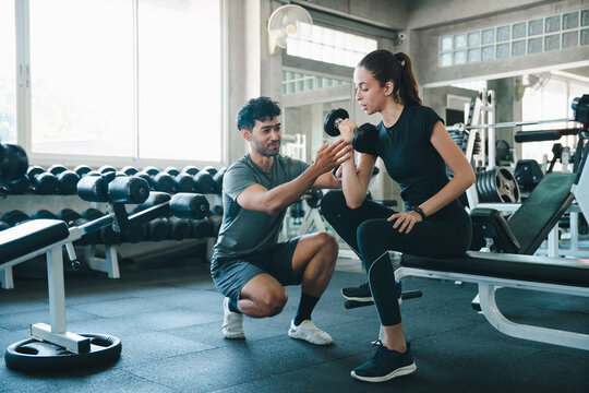 Personal trainer woman dumbbell gym fitness workout exercise strength training coaching athlete guidance on seated arm curl with focused expression in modern weight room