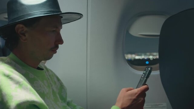 Stylish male traveler wearing leather fedora hat and bright green floral sweater sits in airplane cabin using smartphone while city lights shimmer through oval window during evening flight approach.