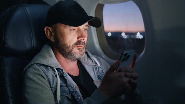 Airplane cabin interior with middle-aged man in black cap and denim jacket using smartphone as aircraft prepares for takeoff. Twilight sky visible through window with city lights bokeh in background.