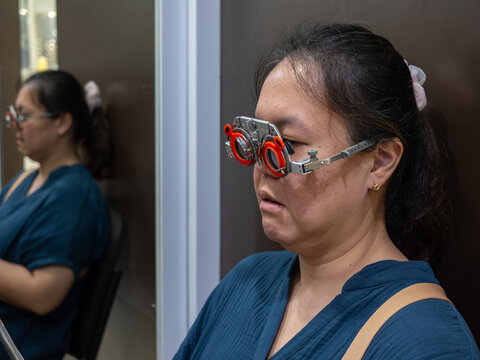A woman undergoes a comprehensive eye examination at an optometry clinic, wearing a metal trial frame (phoropter) with interchangeable lenses to determine her prescription.