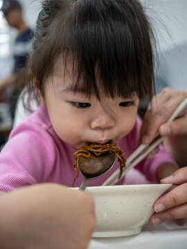 Close-up of an Asian toddler girl eating noodles with a spoon, assisted by adult hands at a casual restaurant.