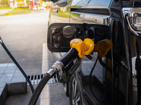Close-up of a yellow fuel nozzle inserted into the open fuel tank of a black minivan at a gas station. The car is being refueled outdoors, with reflections visible on the glossy vehicle surface.