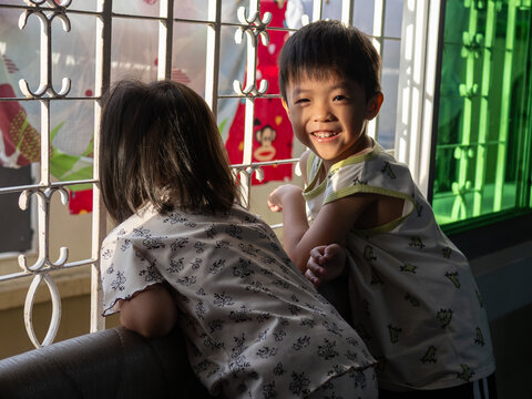 Two young children playing by a sunlit window, with a smiling boy looking at the camera.