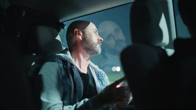 Middle-aged bearded man wearing denim jacket sits in backseat of car at night, using smartphone while gazing out window with city lights reflecting on glass creating moody cinematic atmosphere.