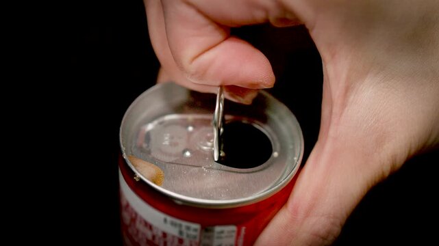 A close-up of a hand opening a metal can by pulling the tab against a dark background in a refreshing beverage scene.