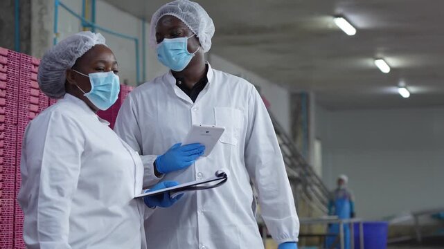 Factory quality control supervisors discussing production data in a food processing plant, Professional team in sanitary gear reviewing inspection results on tablet and clipboard