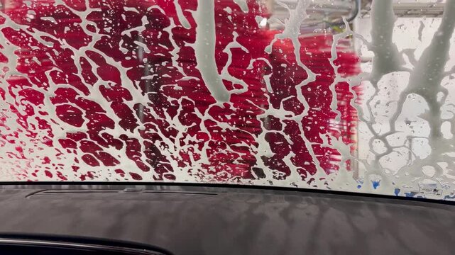 Interior dashboard POV shows swirling soapy suds and vibrant foam patterns on windshield as mechanical brushes move past in a dramatic, close-up car wash with vivid lighting.