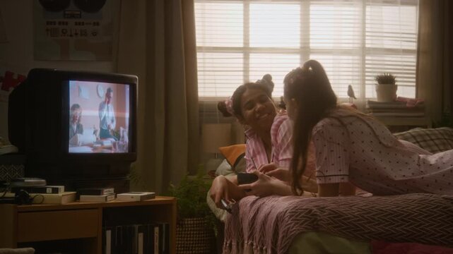 African American teenager and Caucasian girl friend lying on bed in pajamas, watching retro CRT television and eating snacks. Ideal for nostalgic youth marketing and lifestyle campaigns