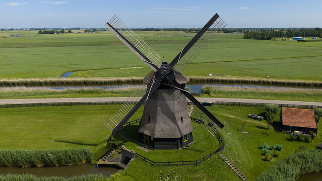 Aerial view of De Museummolen, a historic thatched windmill surrounded by lush green polder fields, canals, and a small outbuilding in Schermerhorn, Noord-Holland, Netherlands.