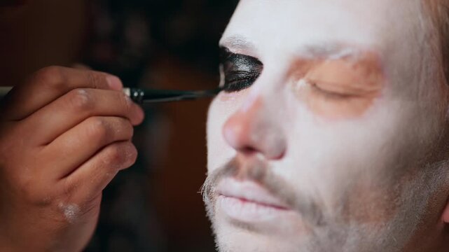 Professional makeup artist applies black eye shadow on man with white face paint for Day of the Dead or Halloween costume. Close-up shows detailed brush work around eye area.
