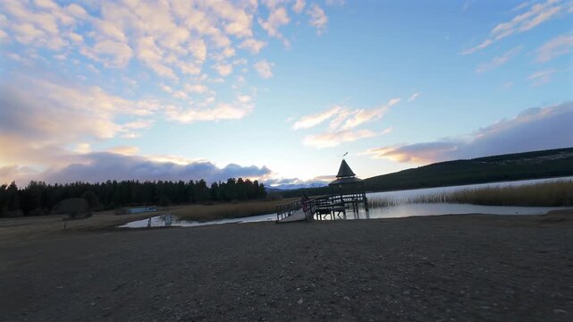 People walking along a wooden dock toward a gazebo at Laguna La Zeta, Argentina. The soft sunset light illuminates the lake and distant mountains.