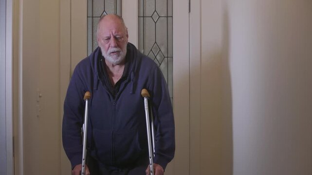 Elderly man with an injured leg slowly walks forward on crutches in a dim hallway, conveying pain and struggle.