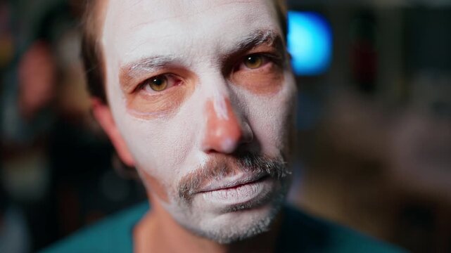 Close-up portrait of middle-aged man with white face paint makeup application for Day of the Dead or Halloween celebration. Intense gaze with dramatic lighting and blurred background.