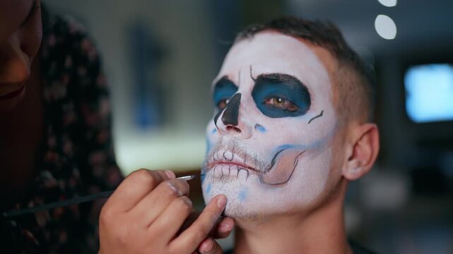 Professional makeup artist carefully applies traditional Day of Dead or Halloween skull face paint on young man using brush. White base with black and blue details creates dramatic sugar skull effect.