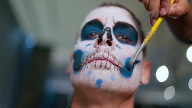 Artist applies traditional Day of the Dead skull makeup on man using yellow brush. White base with black eye sockets and blue accents creates detailed calavera design for celebration.