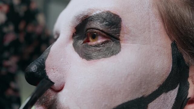 Close-up of artist applying traditional Day of the Dead or Halloween skull face paint. Brush adds black details around nose on white base with dark eye sockets and jawline design.